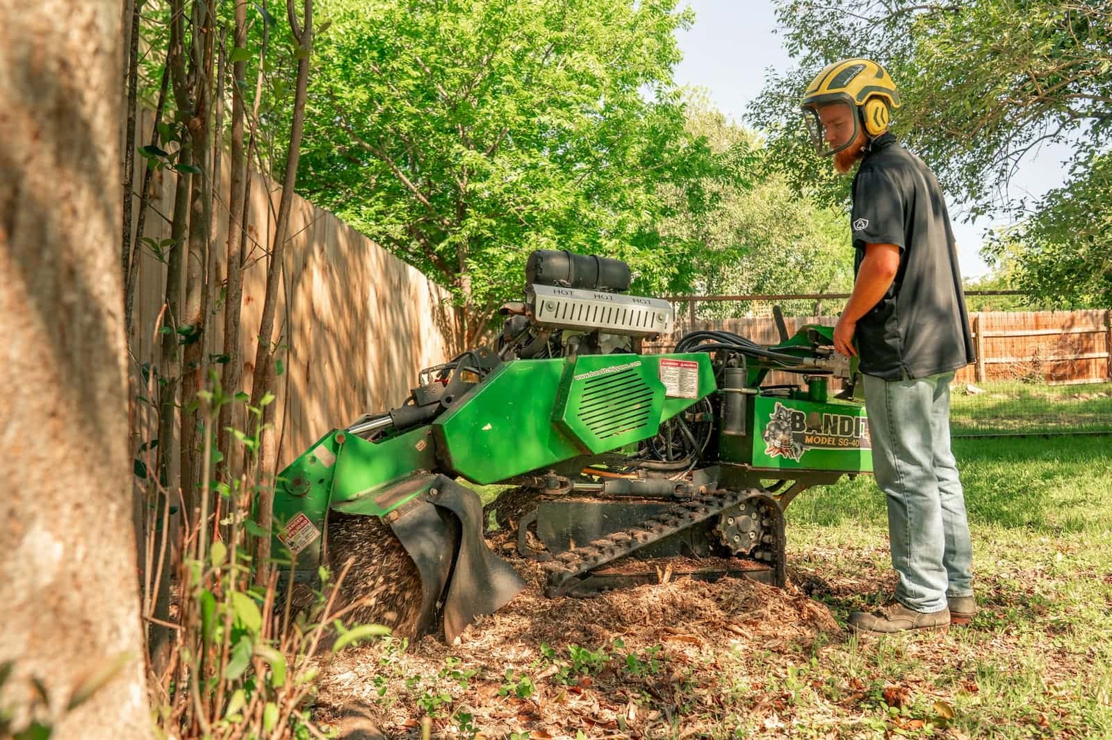 Worker in ear protection running a tracked stump grinder beside a fence in a backyard